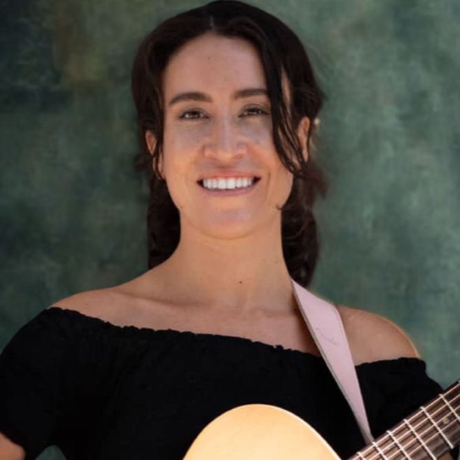 A woman with black hair and tanned skin smiling with a pink guitar strap over the shoulder with the top half of a guitar visible